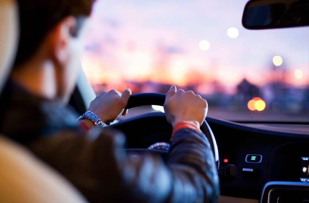 Person driving a car at dusk, holding the steering wheel with both hands as city lights blur in the background.
