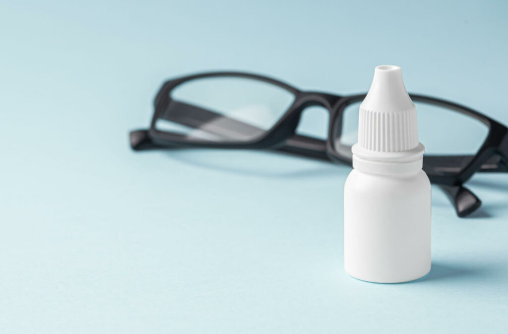 Eye drops bottle placed in front of a pair of glasses on a light background.