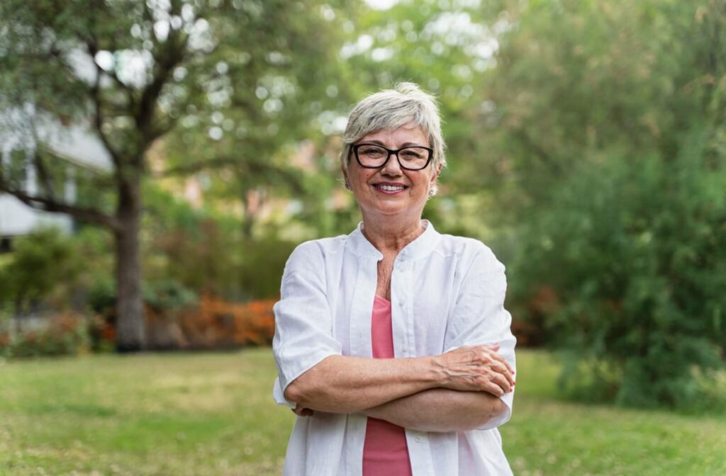 A smiling senior woman with short grey hair and black-framed glasses standing with arms crossed in a lush green park.